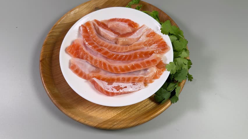 Salted salmon bellies strips without skin on the white dish on wooden stand with fresh cilantro twigs on a gray surface, top view 
