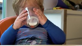 Two-year-old cute blond boy sits on a highchair with a table and drinks milk from a glass
