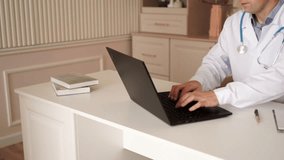 Male doctor wearing uniform with stethoscope and working on laptop in medical office. looking at laptop screen, taking notes in medical journal, filling documents, consulting patient online. Close up