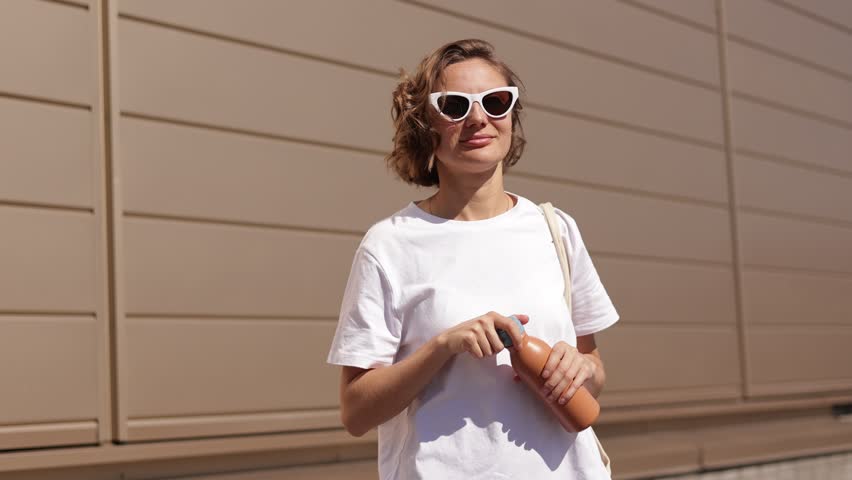 Cheerful millennial young woman opens a reusable metal bottle and drinks fresh water on sunny summer day outdoors on modern building wall background. Zero waste, eco-friendly, no plastic concept. Royalty-Free Stock Footage #3428673055
