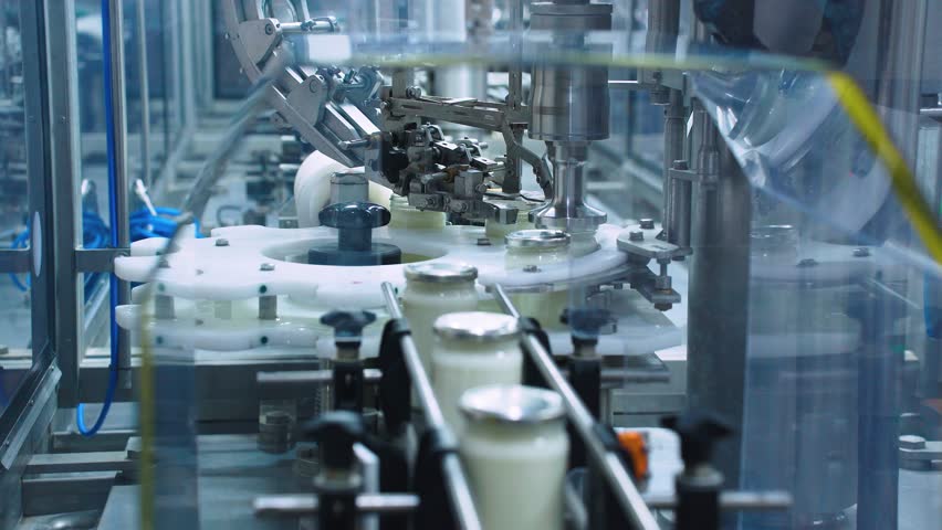 A packaging machine at a dairy factory automatically screws caps onto glass jars of milk or yogurt. Packing and bottling of dairy products. Vacuum modern packaging of food products Royalty-Free Stock Footage #3428736731