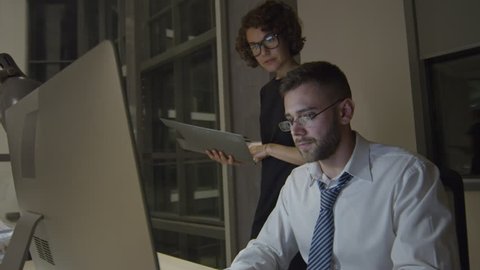 Colleagues working in modern glass office in the night: businessman typing on computer and speaking with female colleague