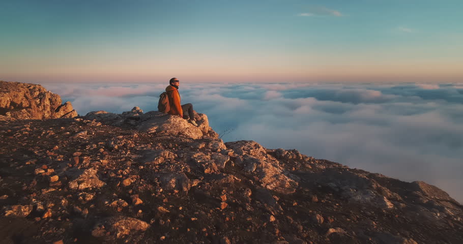 Young happy girl hiker with backpack is sitting at mountain top above the clouds and enjoys golden sunset and great view