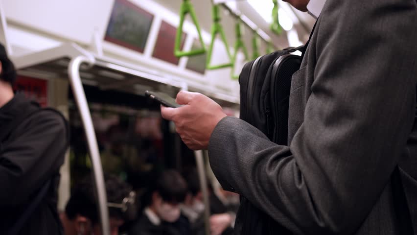 A Japanese businessman in the subway, engrossed in his smartphone, seamlessly blends business with the urban rhythm of the city.