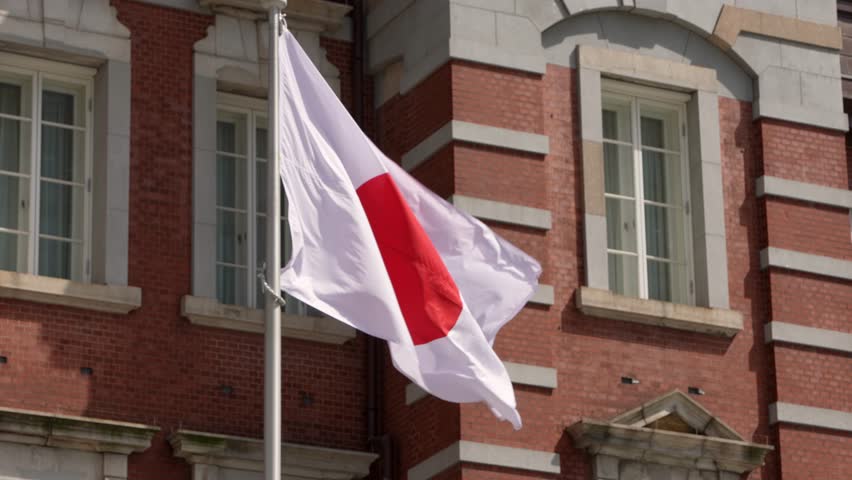 Slowmotion of a Japanese flag waving in wind at traditional building of train station. Japan banner blowing, soft silk. Cloth fabric texture ensign background. Use it for national day country concept.