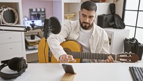 Bearded man playing guitar in a well-equipped home music studio with microphone and headphones.