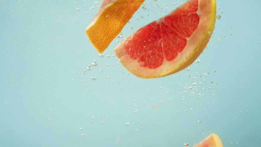 Pieces of grapefruit one by one fall into the water on a blue background. Slow motion background video with citrus in liquid. High quality 4k footage