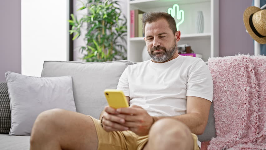 A middle-aged man with grey hair smiles while using a smartphone on a sofa at home, representing a casual lifestyle. Royalty-Free Stock Footage #3428931871