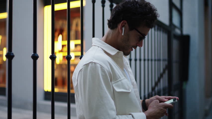 Introspective young man in his 30s, with curly hair and glasses, engrossed in smartphone whila standing outdoors, urban alley with warm lighting, depicting concentration and modern connectivity Royalty-Free Stock Footage #3428966161