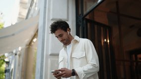Young adult male in headphones wearing white shirt and jacket, man texting message on his smartphone while standing on city street at building entrance, exuding relaxed urban vibe, during spring - Powered by Shutterstock - Get 15% off with code: PIKWIZARD15