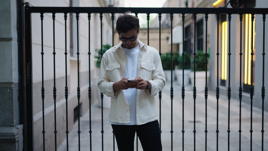 Hispanic young man in 30s, with curly hair and glasses, engrossed in his smartphone while scrolling social media web, urban alley with warm lighting, depicting concentration and modern connectivity Royalty-Free Stock Footage #3428967657