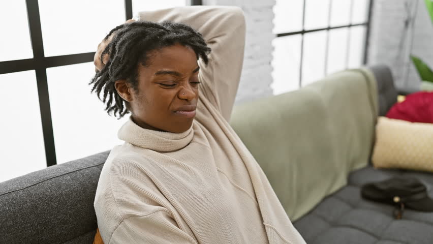 Thoughtful young black woman with dreadlocks, sitting on the sofa at home, wrestling with a question. her doubtful expression reveals a pensive mood, curious and unsure in concept. Royalty-Free Stock Footage #3429011489