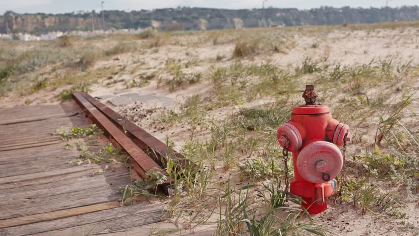 Rusty red fire hydrant on a sandy beach with wooden pathway and grass. Concept of safety, preparation, and contrast between man-made and natural environments.