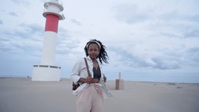 Video beautiful young African American woman looking smiling camera. People enjoying sightseeing on summer vacation. Happy girl posing on coast with sea lighthouse in background. Weekend getaway.