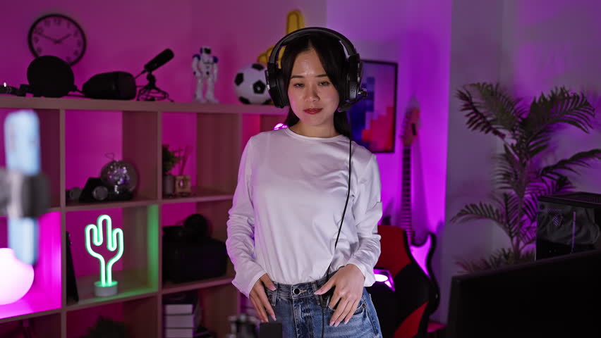 A cheerful young asian woman with headphones poses in a vibrant gaming room at night, exemplifying modern home entertainment.