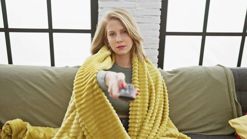 Blonde woman with remote control sitting on sofa at home, looking at viewer.