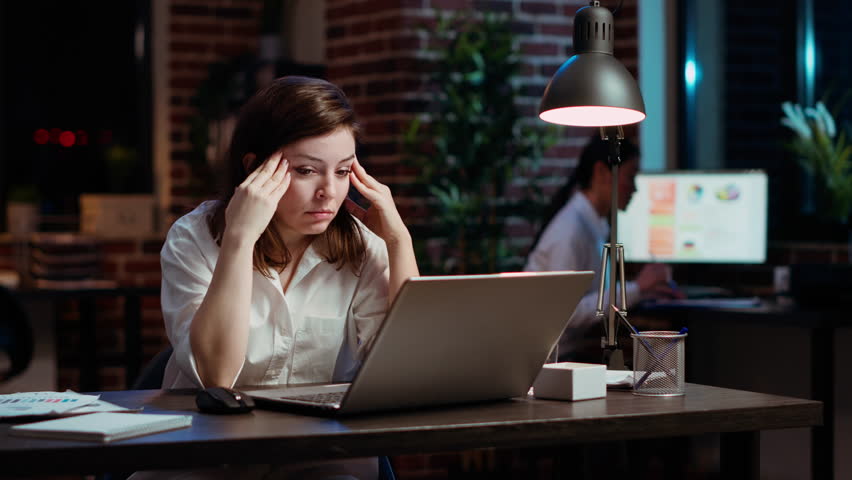 Tired employee struggling to remain focused at workspace desk, feeling overworked and waiting to go home. Bored businesswoman sighing in office while working overnight, camera B