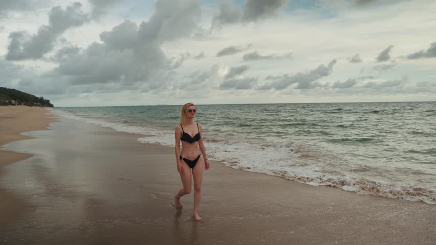 Beautiful woman walks along sandy tropical beach in black bikini enjoying a tropical vacation