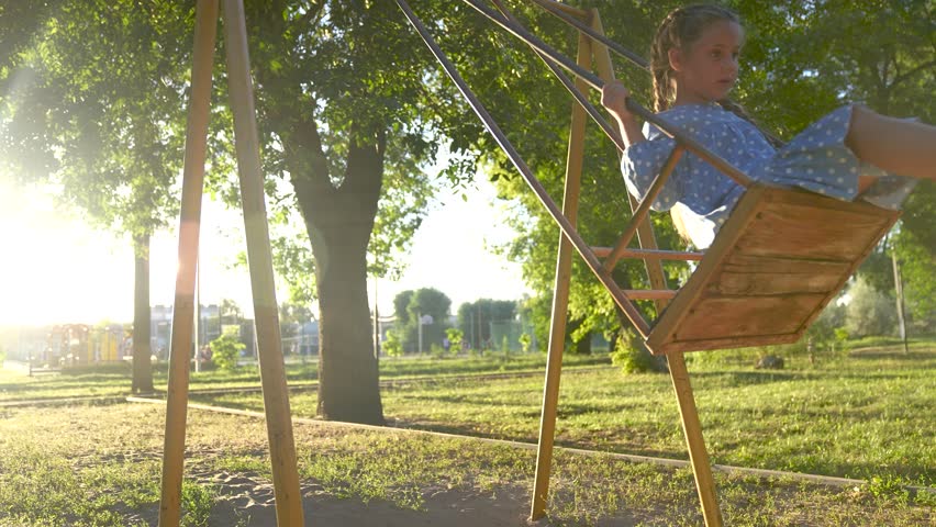 school girl swings on swing. concept of a lifestyle happy childhood and loving family. little child in blue dress swings on a swing on the playground, sunset and park in the background
