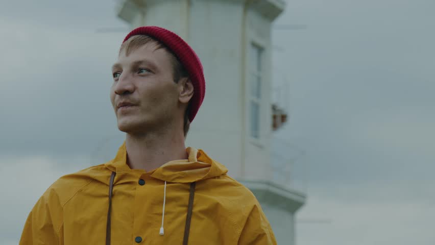 Portrait: A young man in a red hat and a yellow jacket stands against the background of a lighthouse, looking into the distance. The lighthouse keeper. A traveler enjoys nature, Slow Motion, 4K