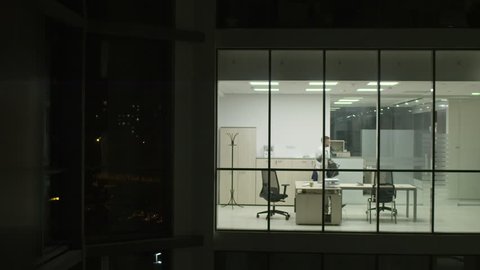 View from outside of modern glass office of businessman taking briefcase and coat, leaving workplace at night and turning off the light