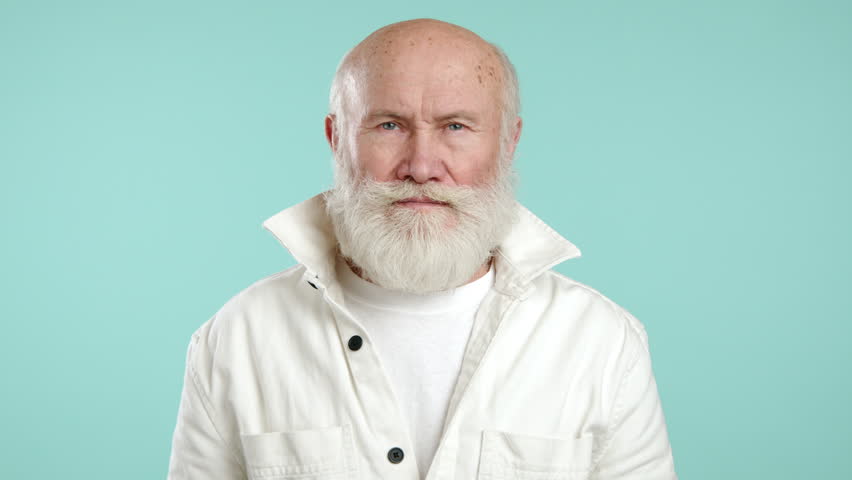 A bashful elderly man in a youthful white jacket covers his face with his hand against a turquoise backdrop, portraying a moment of shyness or embarrassment. Camera 8K RAW.  Royalty-Free Stock Footage #3429283093