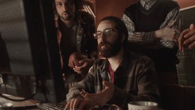 Medium shot of long haired bearded Middle Eastern geek programmer showing new developed software on computer to multiethnic colleague, working together in dark old-fashioned office with dimmed light