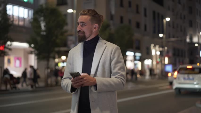 Cheerful caucasian man walking at night in a lively city, using phone to send online messages or looking for a place. Copy space.