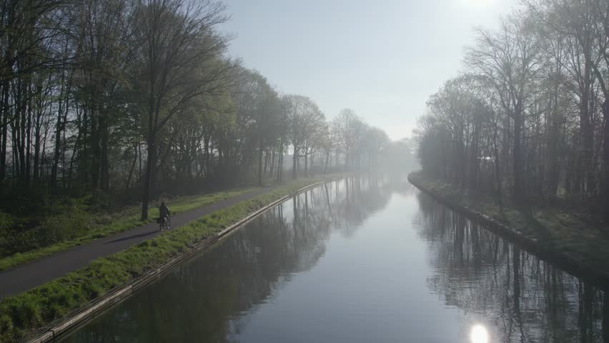 This serene stock footage depicts a solitary figure cycling along a canal path enveloped in the soft caress of morning mist. The tranquility of the scene is palpable as the drone follows the cyclist