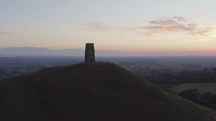 Wide sunset aerial orbiting the Glastonbury Tor set against countryside and colourful skies in Somerset, United Kingdom