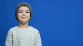 Cute child in hat and gloves laughing and covering his face with his hands on blue background.