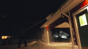 Removing snow from roof with long telescopic snow roof rake, middle age man work with snow removal tool during dark time of day. Rear side view video