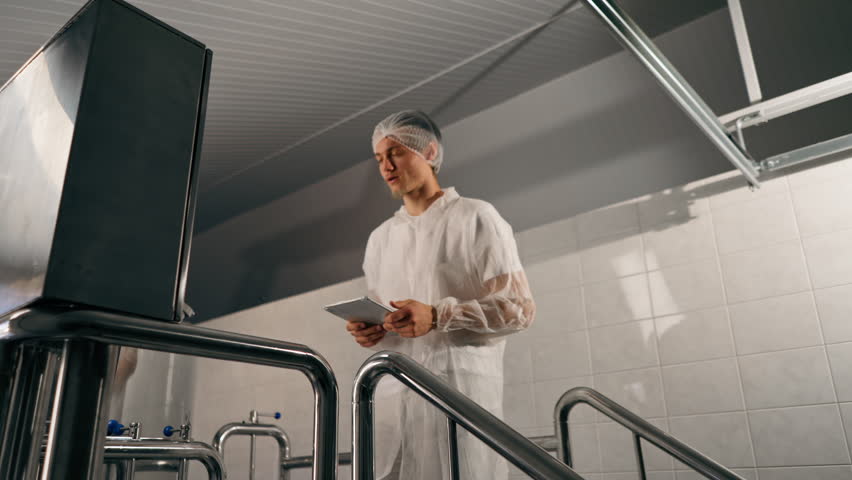 A male technologist in a protective suit with a tablet in his hands checks the production stage of fermentation of a drink at a brewery Royalty-Free Stock Footage #3429642531