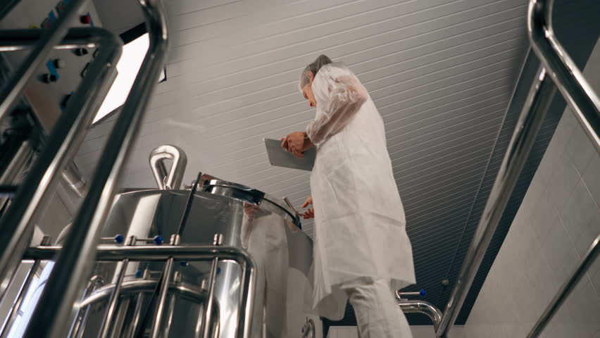 A male technologist in a protective suit with a tablet in his hands checks the production stage of fermentation of a drink at a brewery Royalty-Free Stock Footage #3429647097