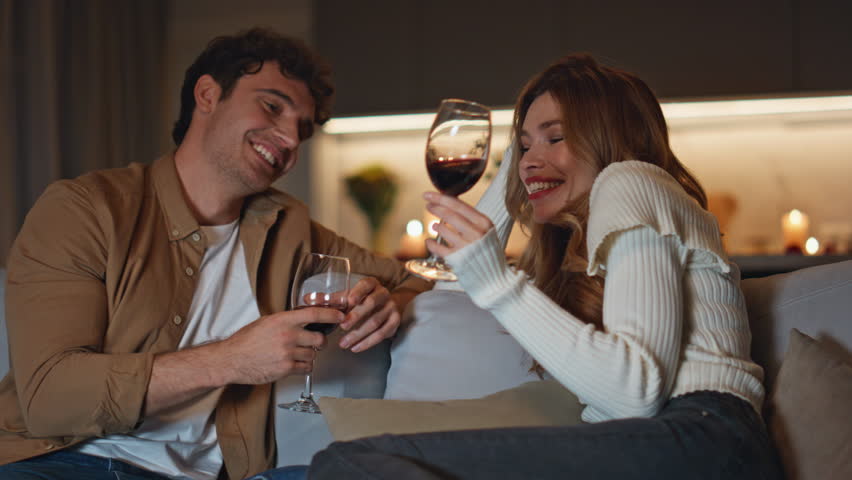Married pair enjoy date on cozy home couch clinking wine glasses close up. Smiling young couple spending romantic evening in candles atmosphere. Happy lovers relaxing together drinking wine indoors.
