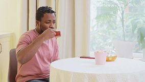 African american guy hipster drinking herbal tea sitting on kitchen at home near window. Serious thoughtful man with dreadlocks. Wering pink t-shirt and jeans. Everyday routine, having meal concept. - Powered by Shutterstock - Get 15% off with code: PIKWIZARD15