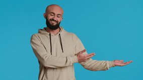 Cheerful man creating ad in front of studio camera, pointing to left or right sides over blue background. Middle eastern person showing commercial or advertisement for marketing campaign. Camera 1. - Powered by Shutterstock - Get 15% off with code: PIKWIZARD15