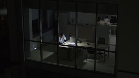 View from outside of modern glass office of tired overworked businessman drinking coffee and rubbing his face in front of computer at night