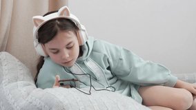 A little relaxed girl lying on the bed with headphones with cat ears listening to music or watching videos. He holds a smartphone in his hands. A teenager on a day off.