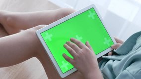 A tablet in the hands of a little girl on her lap with a green screen for advertising applications, games or children's channels. A child in a blue hoodie touches the display with his finger.