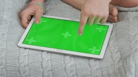 A tablet with a green chrome kay screen lies on a gray knitted plaid, a little girl touches the display with her finger. Bare feet. Advertising an app, video games.