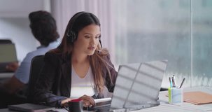 Indian happy woman wearing headphone sitting on chair using laptop talking video call centre indoor office. Focused freelance female agent provide customer support service speaking client at workplace