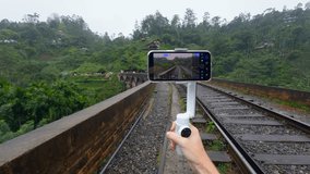 Shooting on tripod phone while traveling. Action. Shooting video on phone with stabilizer while traveling. Shooting bridge with railway in jungle on phone for blog