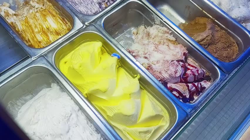Seller scoops a portion of yellow lemon ice cream sundae into a spoon from the refrigerator. Trays with various tastes. Selling food on the street at night
