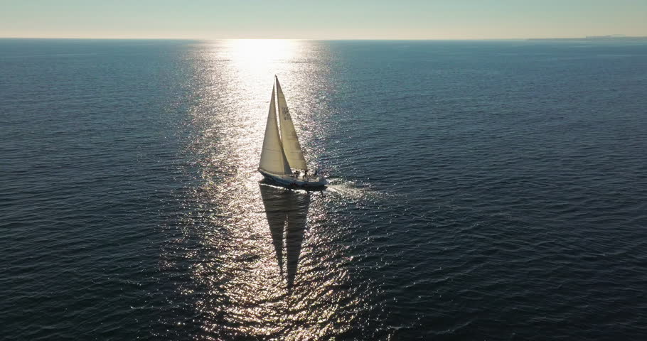 Tracking shot following a yacht into the sunset. Big sailing yacht boat driving on the ocean on a sunny day. Floating sailing yacht with spread sails splitting the sea waves