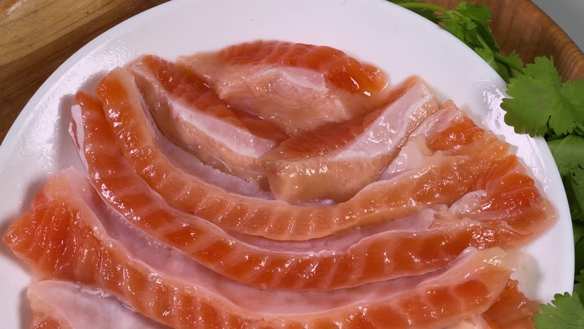 Salted salmon bellies strips without skin on the white dish on wooden stand with fresh cilantro twigs, top view close-up 
