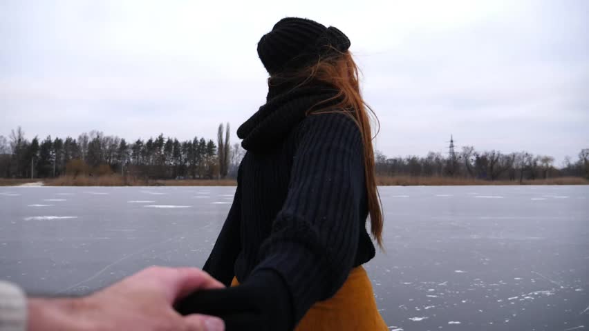 Follow me shot of beautiful red-haired woman holds male hand sliding on figure skate at frozen river. Young girl pulls her boyfriend skating on ice surface. Happy couple having active lifestyle. POV
