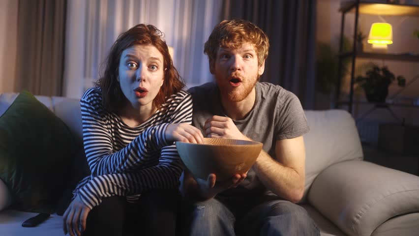 Close up portrait of addicted focused young red haired couple watching interesting TV program film movie and eating popcorn at home Enchanted family cannot take eyes off the screen indoors