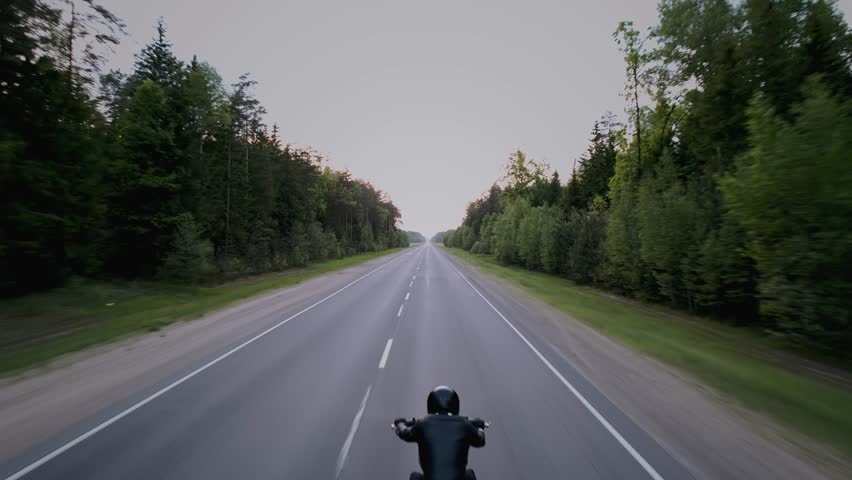 Aerial view of man in black leather jacket and black helmet riding motorcycle along suburban highway. Back view