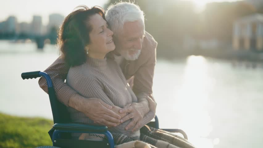 Husband walking with wife in wheelchair. Senior family couple in city park. Old man hugging woman showing love care, they talking smiling. Living with physical disability, long love concept. Royalty-Free Stock Footage #3430085179